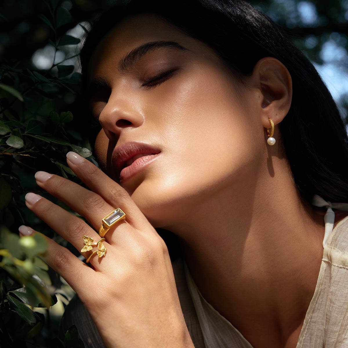 A woman with closed eyes poses outdoors, touching her face. She wears 18k gold plated hoop earrings with pearls and two gold rings—an adjustable ring with a rectangular stone and the Freedom to Soar Adjustable Butterfly Double Butterfly Ring. Sunlight softly illuminates her.