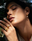 A woman with closed eyes poses outdoors, touching her face. She wears 18k gold plated hoop earrings with pearls and two gold rings—an adjustable ring with a rectangular stone and the Freedom to Soar Adjustable Butterfly Double Butterfly Ring. Sunlight softly illuminates her.