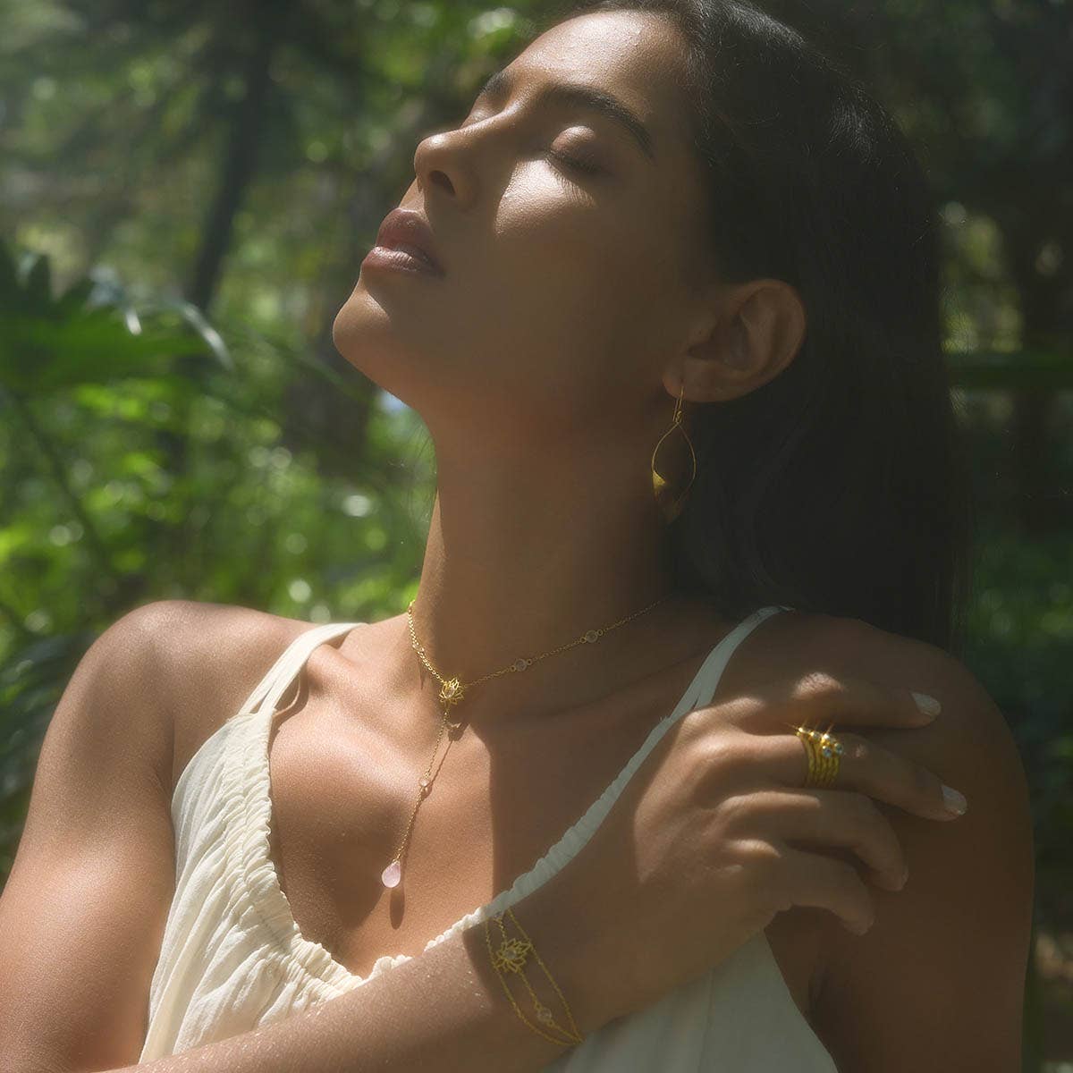 A woman in a white dress stands outdoors with eyes closed, basking in sunlight. She wears gold earrings and the Transformed by Love Lotus Lariat Necklace. Green foliage forms a blurred background.