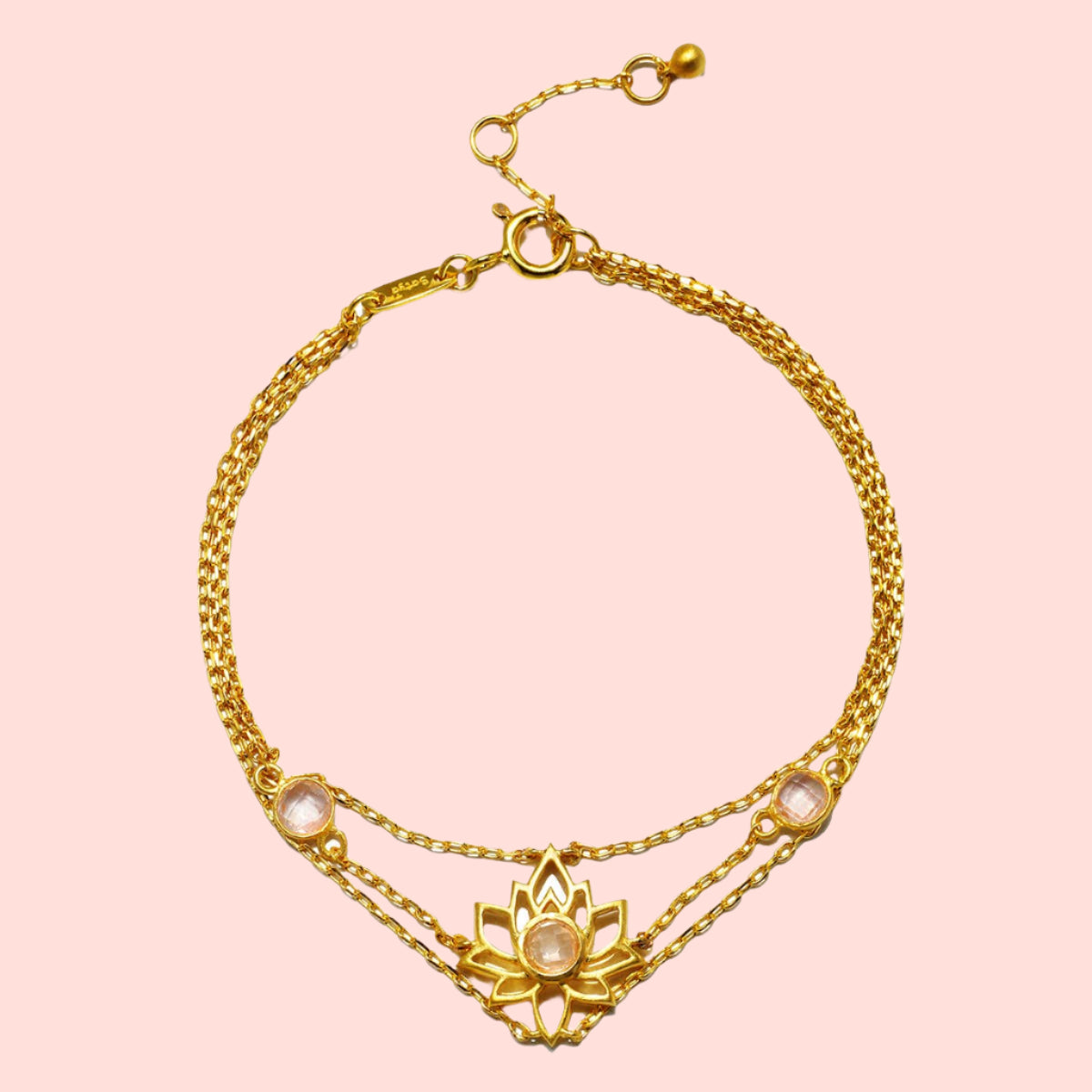 The Transformed by Love Lotus Bracelet is an 18k gold piece with a central lotus flower and a pink gemstone, flanked by two smaller pink stones and an adjustable clasp—ideal for lotus jewelry lovers.