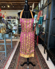 A black mannequin is adorned with a Fancy & Adorned Handmade Sari Apron that showcases a vibrant gold and pink floral design. This luxurious apron features a golden yellow hem and is displayed in a beautifully decorated store with shelves containing folded fabrics and other items in the background.