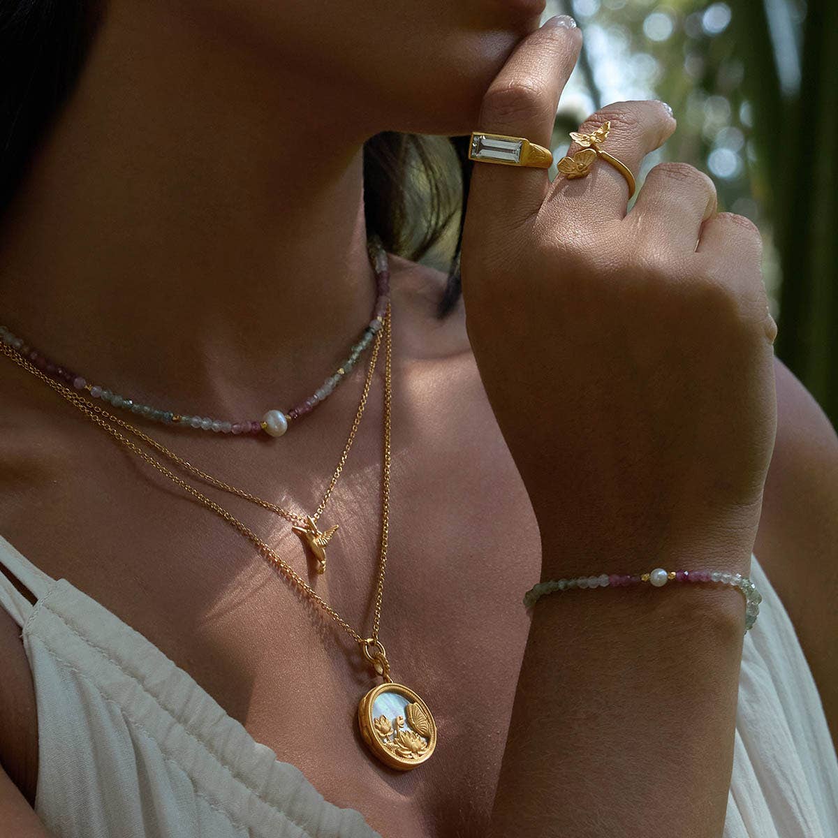 A woman wears the Take Flight Butterfly Pearl Locket Necklace 18” with gold rings and a beaded bracelet, styled over a light-colored sleeveless top in natural light.