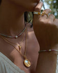 A woman wears the Take Flight Butterfly Pearl Locket Necklace 18” with gold rings and a beaded bracelet, styled over a light-colored sleeveless top in natural light.