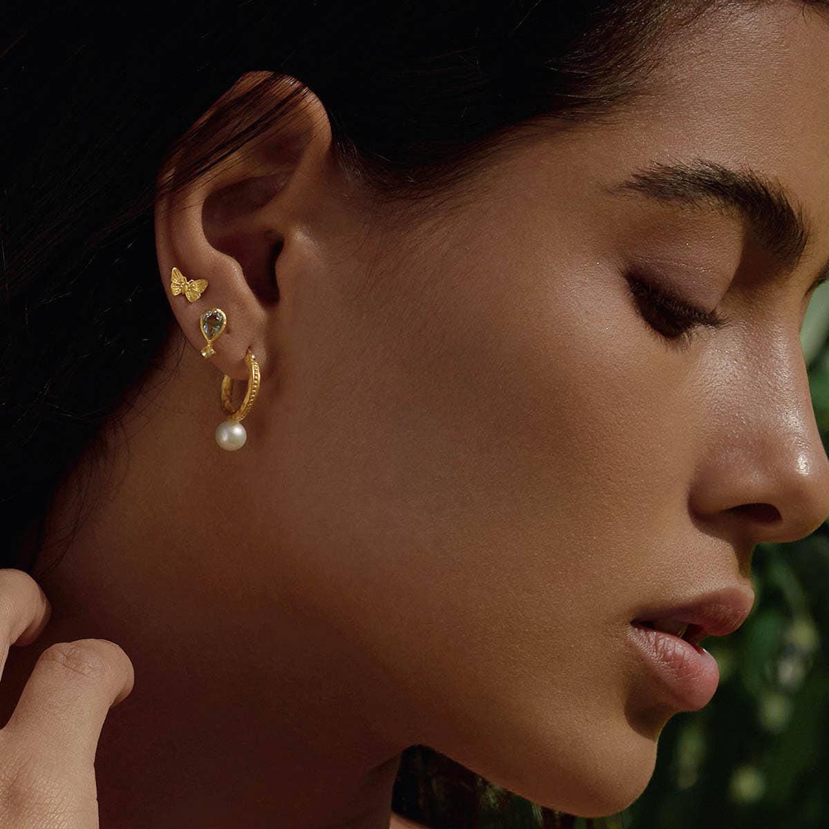 A woman with smooth skin wears the Freedom to Soar Butterfly Stud Earrings, a small crystal stud, and a gold hoop with a dangling pearl, gazing downward serenely against a softly blurred background.
