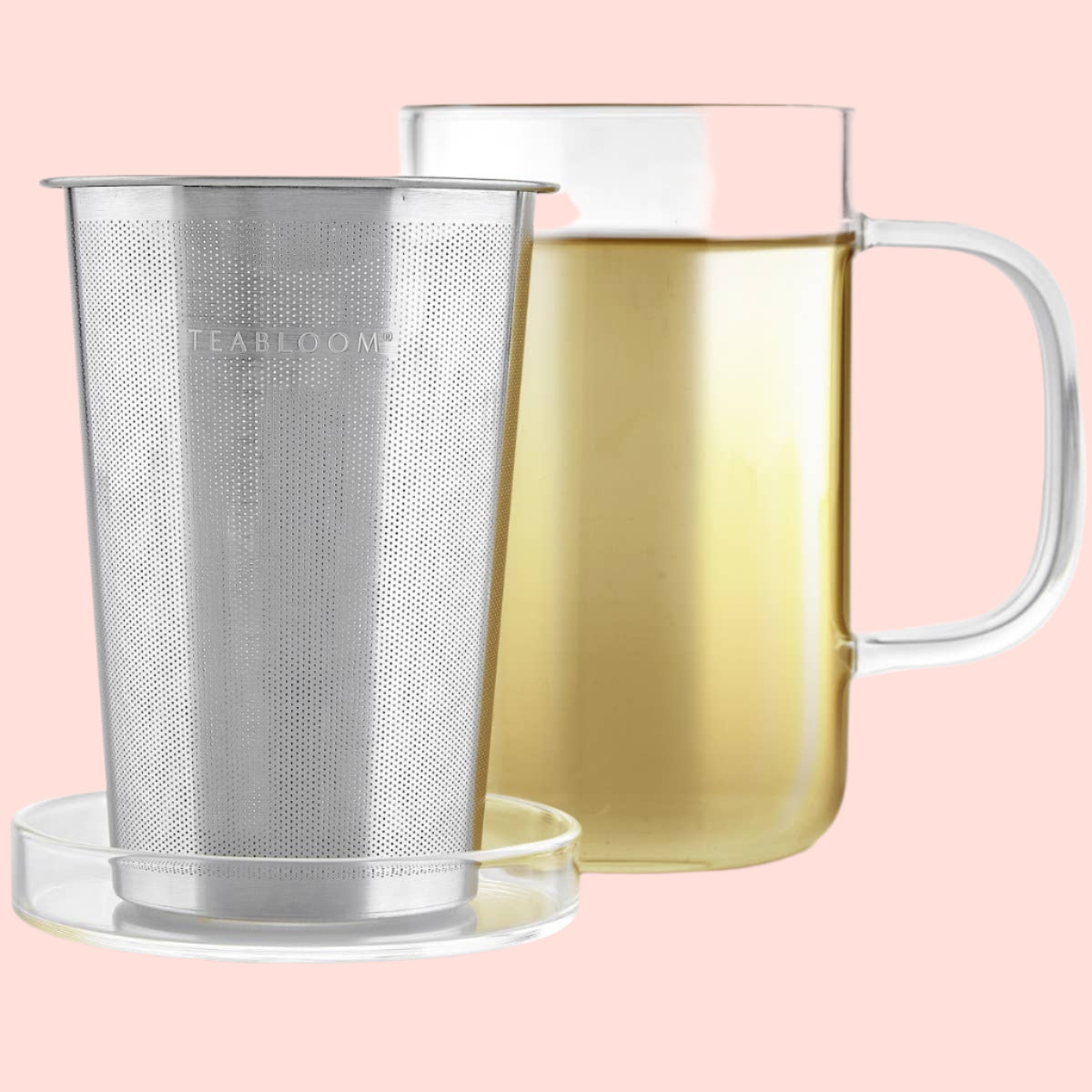 A Teabloom Balance Single-Serve Tea Maker (12 oz) with fine holes for optimal brewing rests on a clear glass coaster, in front of a borosilicate glass mug filled with light-colored tea. "TEABLOOM" is printed on the cylindrical stainless steel infuser.