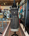 A black dress with a white pattern is displayed on a mannequin in a boutique. The dress features a halter neckline and a decorative white border at the hem. Nearby, Fancy & Adorned Handmade Sari Aprons hang elegantly amidst the rugs, tables, and shelves filled with various items and colorful fabrics.