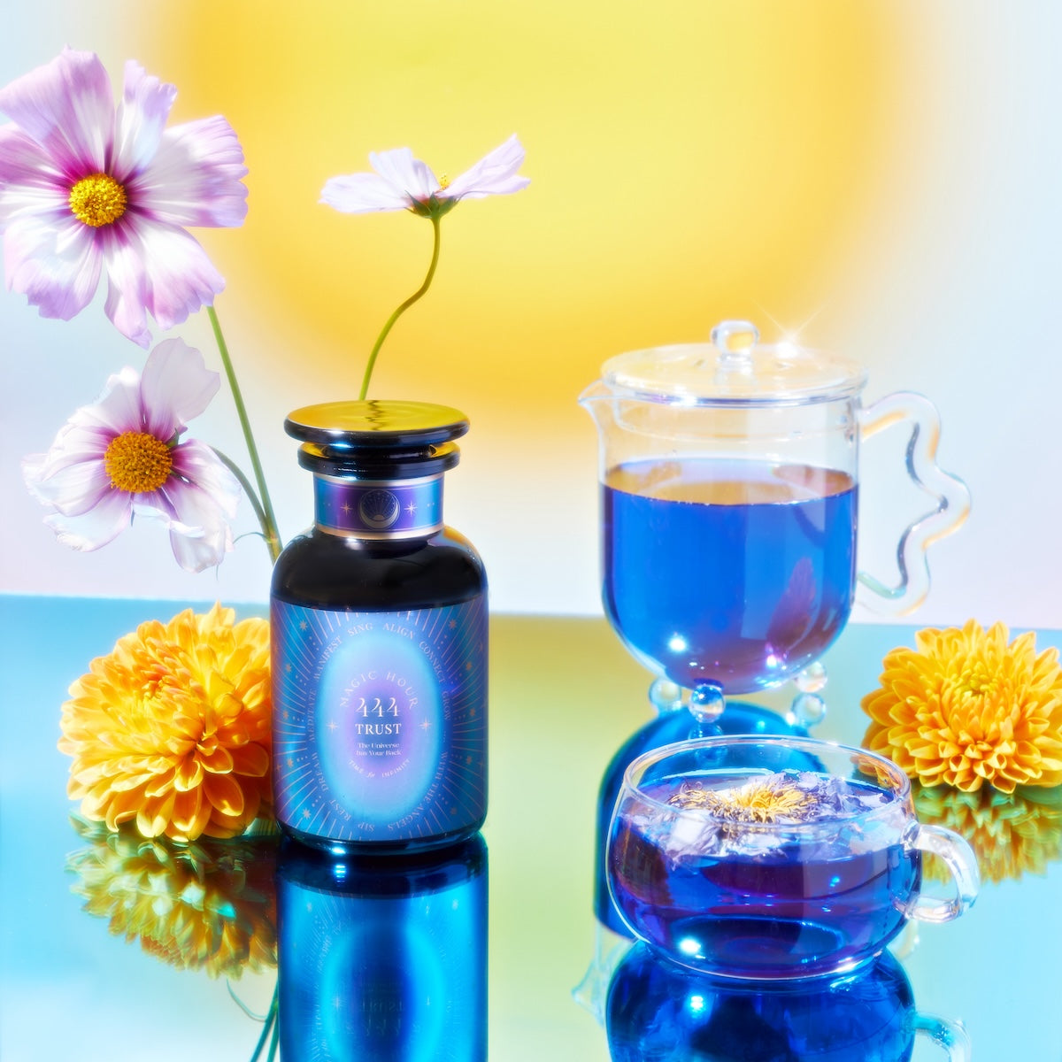 The vibrant still life presents a Petite Apothecary Jar from the School of Magic Tea Subscription Box beside a glass teapot and cup filled with blue wellness tea. Yellow chrysanthemums and pink cosmos enhance the scene, set against a colorful gradient backdrop, inviting mindfulness.