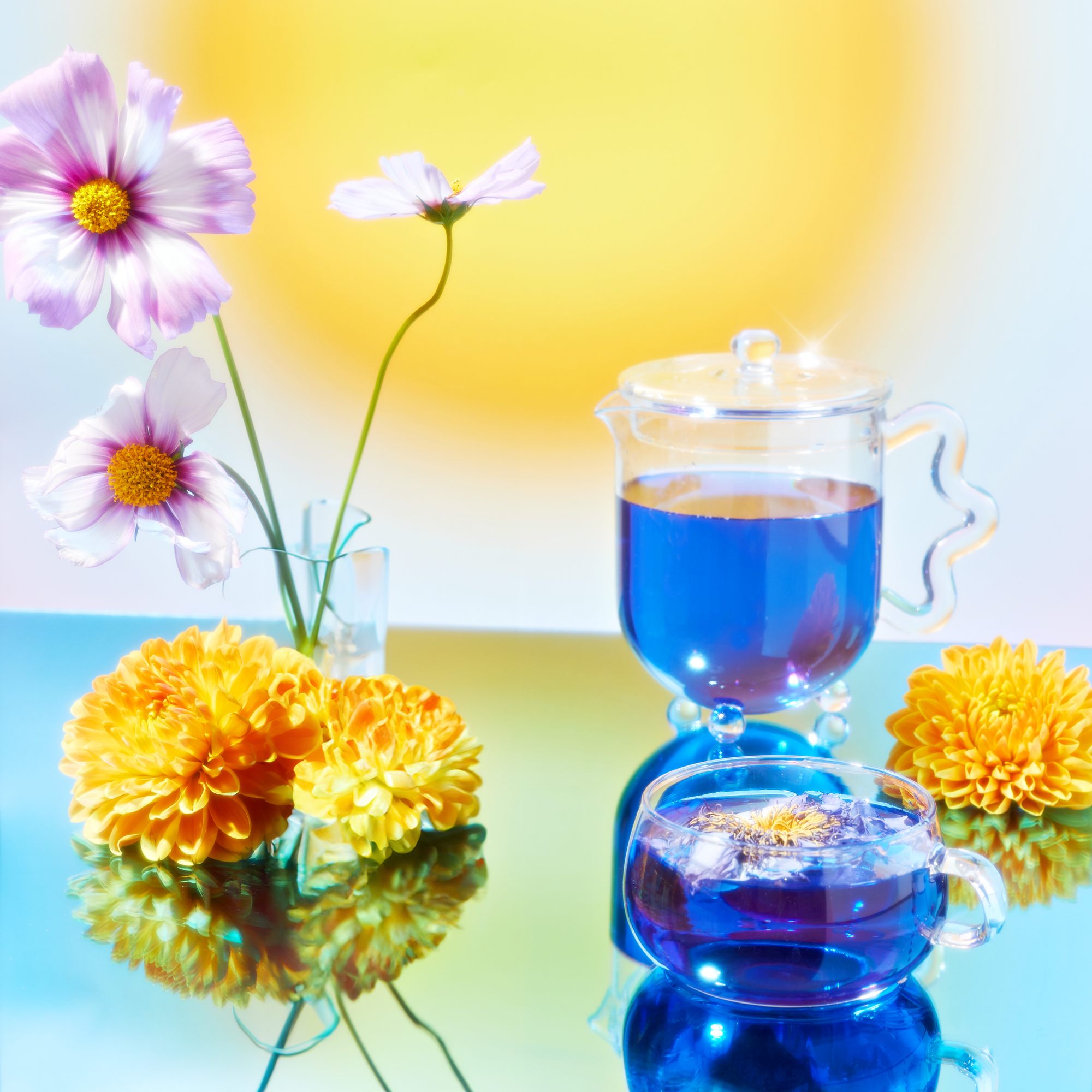 A teapot and cup with vibrant blue wellness tea from the School of Magic Tea Subscription Box - Traveler Jar sit on a reflective surface, evoking spiritual connection. Pink cosmos and yellow chrysanthemums are artfully arranged, with a sunny, blurred backdrop enhancing the serene composition.