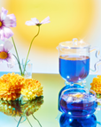 A teapot and cup with vibrant blue wellness tea from the School of Magic Tea Subscription Box - Traveler Jar sit on a reflective surface, evoking spiritual connection. Pink cosmos and yellow chrysanthemums are artfully arranged, with a sunny, blurred backdrop enhancing the serene composition.