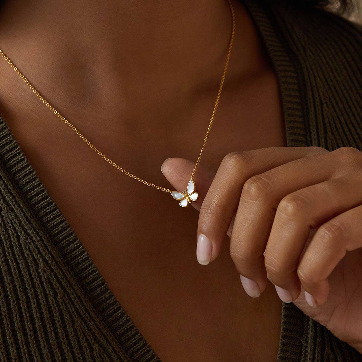 A woman wearing a ribbed olive-green top gently holds the Renewed Hope Pearl Butterfly Necklace, its 18k gold plated chain shimmering and highlighting the delicate mother of pearl butterfly pendant between her fingers.