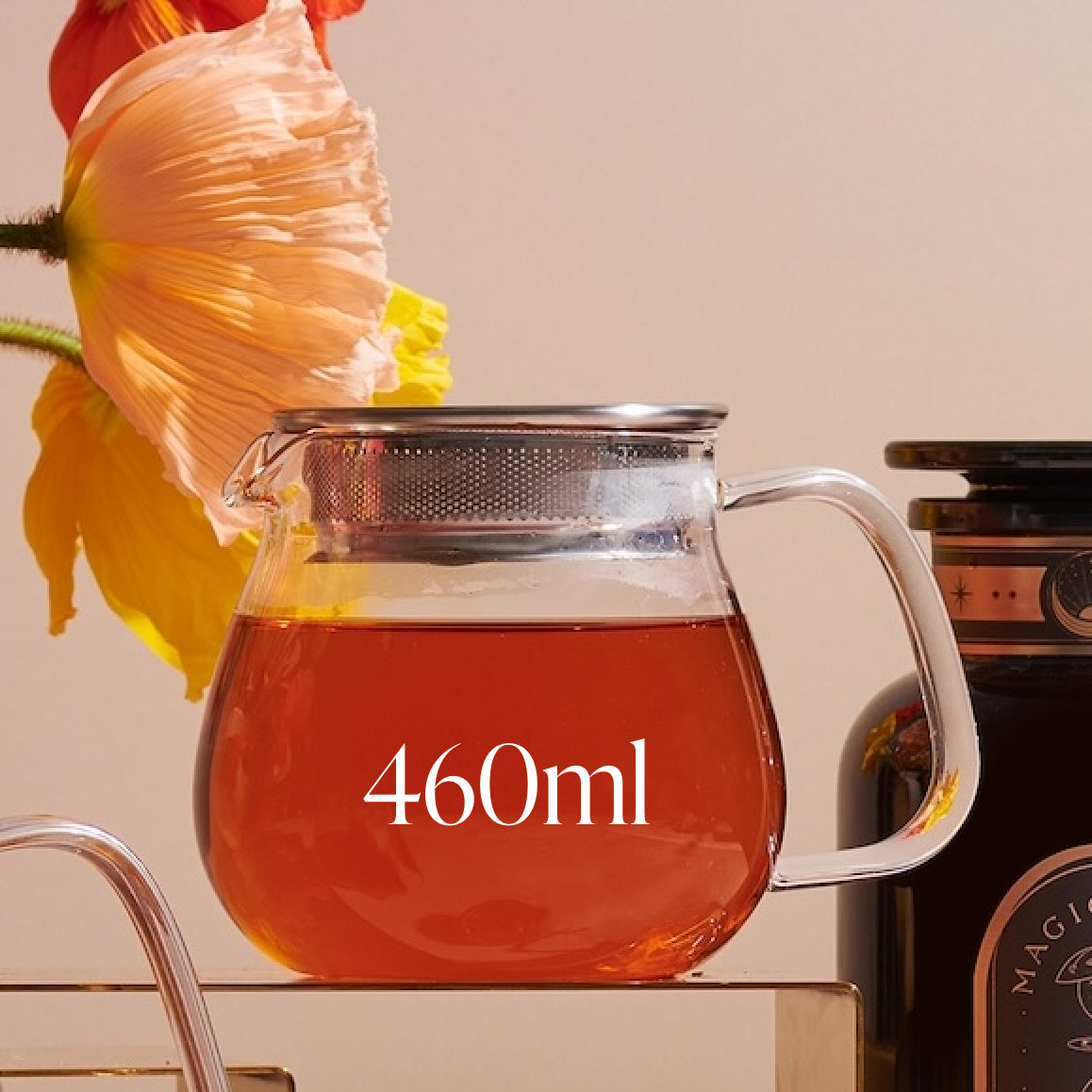 A glass Kinto One Touch Teapot filled with loose leaf tea, labeled "460ml," sits on a clear surface. The teapot has a metal lid with a built-in strainer. Behind the teapot, there is a blooming flower and an out-of-focus dark jar with an organic tea label on it.