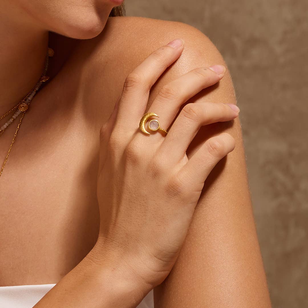 A woman gently rests her hand on her shoulder, highlighting the Cradled in Light Moonstone Ring—gold plated with a crescent moon design and round white stone—set against a neutral brown background.