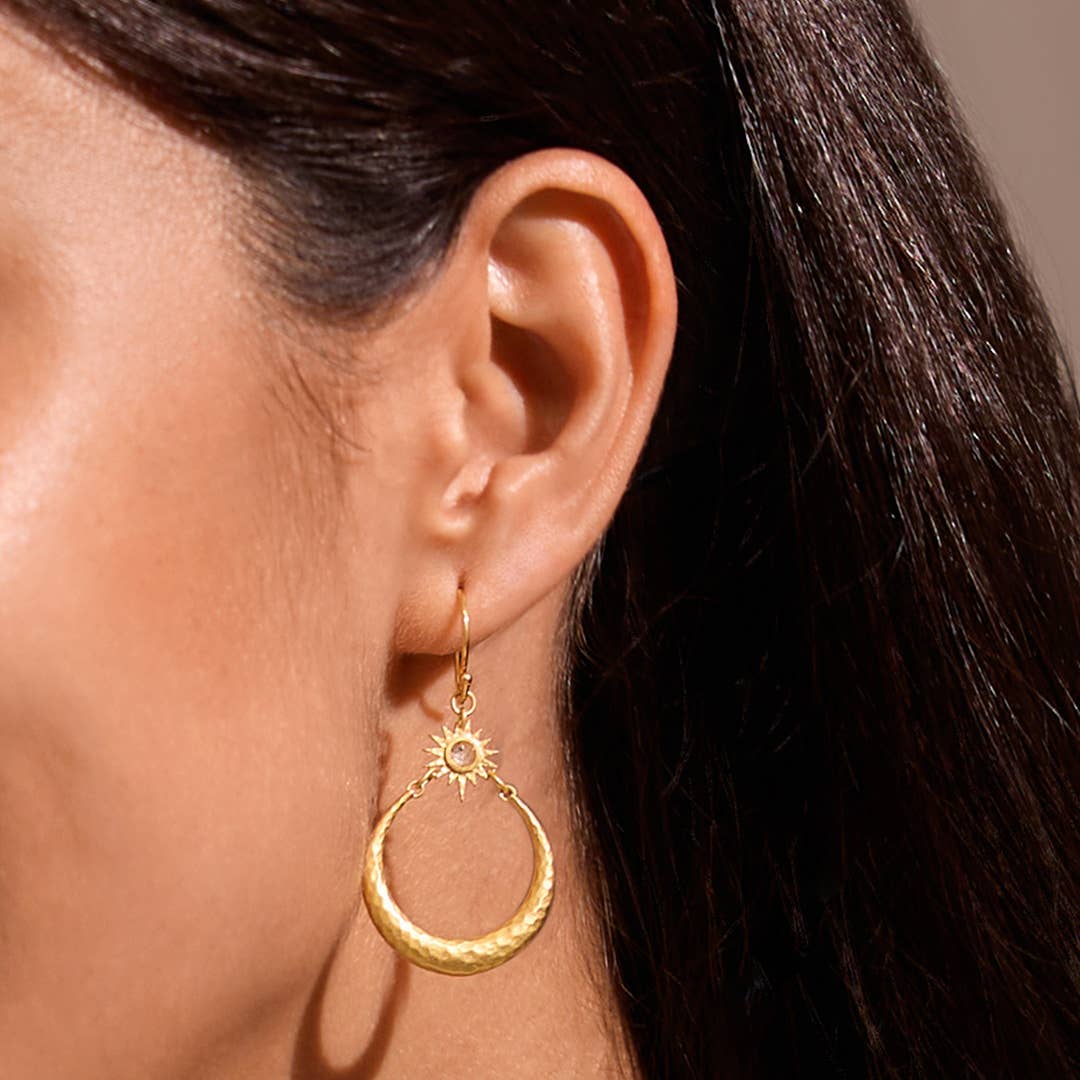 A close-up of a woman’s ear wearing the Stellar Sisterhood Moonstone Hoops—gold crescent moon earrings with a small sun design above the crescent. Her straight, dark hair is tucked behind her ear.
