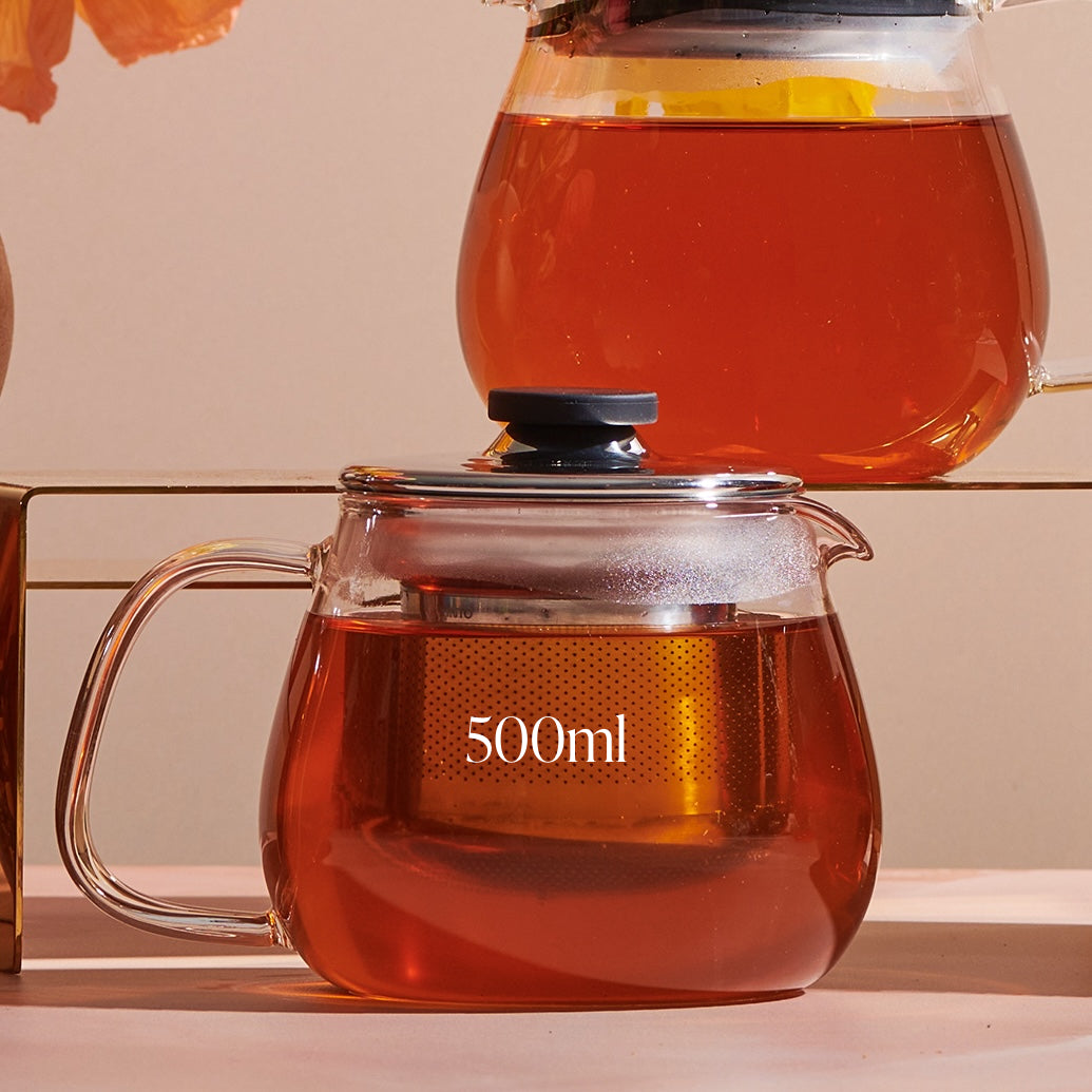 A clear glass Kinto Stainless Steel Teapot from Magic Hour, with a black lid, holding 500ml of amber-colored loose leaf tea, sits on a reflective surface. Another similar teapot is partially visible in the background, positioned on an elevated surface. Both teapots feature metal infusers.