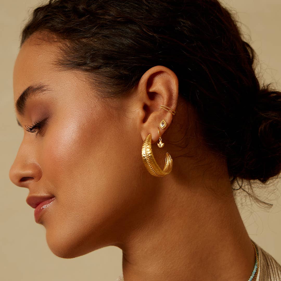 A woman with curly dark hair in a low bun wears several gold earrings, including a large textured hoop, small studs, and Inner Light Blue Topaz Pear Stud Earrings—symbolizing new beginnings—set against a neutral background. Her skin glows softly.