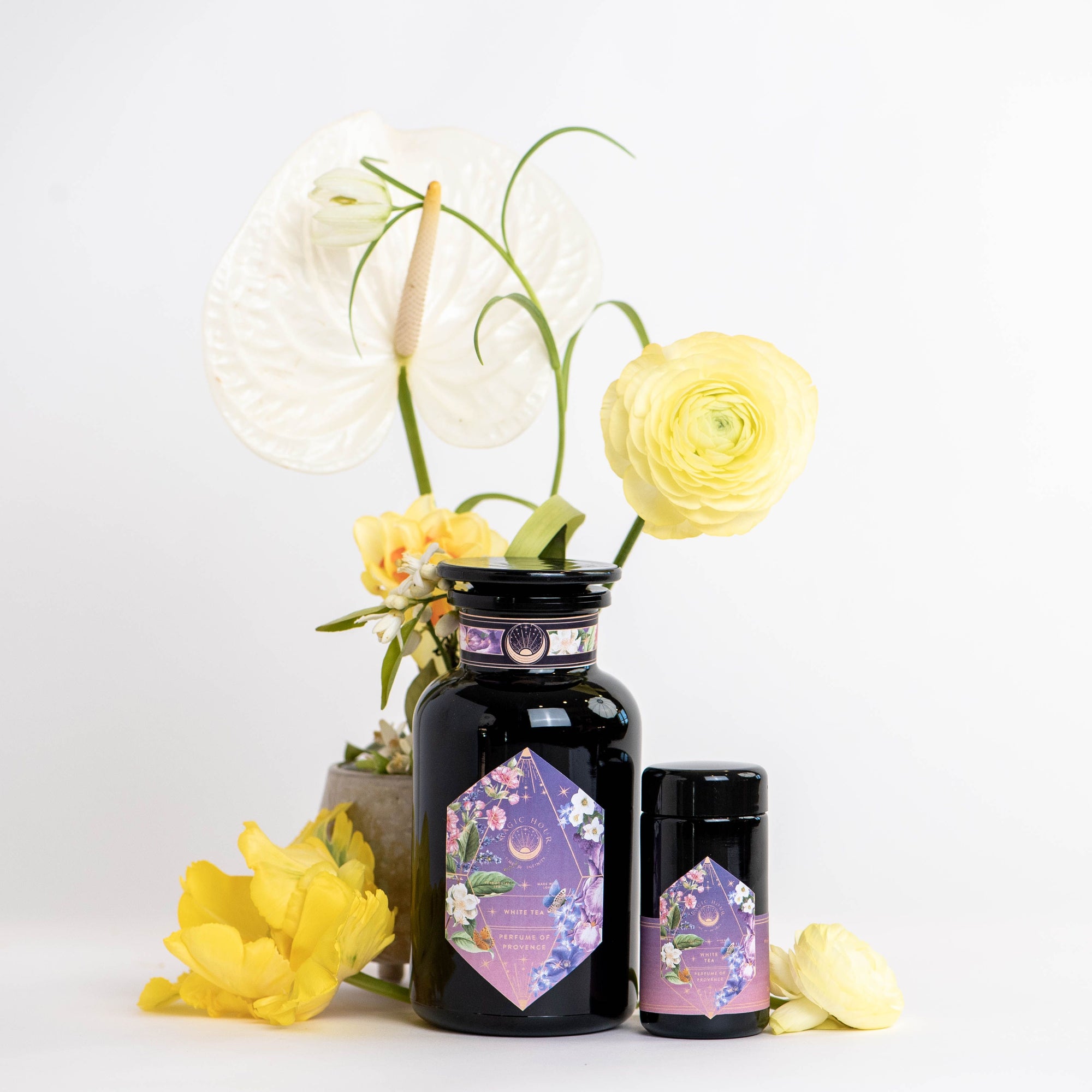 Two dark glass jars with hexagonal purple floral labels for "Perfumes of Provence: Lavender Bouquet Tea for Calm Moods and Beautiful Skin" are placed beside yellow and white flowers, including a large white anthurium, against a white background.