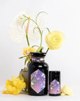 Two dark glass jars with hexagonal purple floral labels for "Perfumes of Provence: Lavender Bouquet Tea for Calm Moods and Beautiful Skin" are placed beside yellow and white flowers, including a large white anthurium, against a white background.
