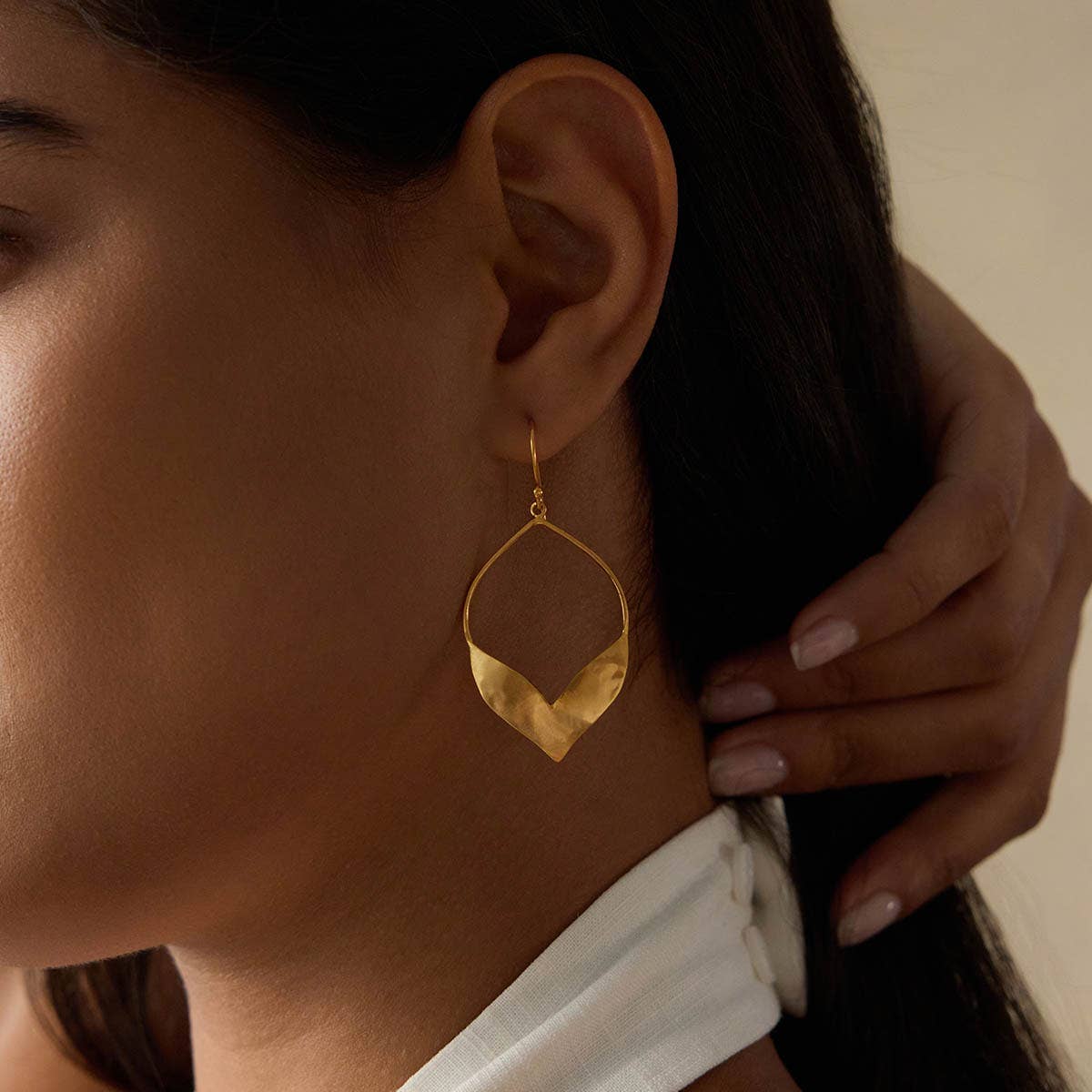 Close-up of a woman wearing Fluttering Petals Gold Earrings, large and modern with a curved leaf design in 18kt gold plating. She touches her neck and wears a white top with visible back button details.