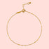 The 9" Shimmering chain anklet + 1" extender chain features delicate 18kt gold plating, tiny textured beads spaced along a shimmering chain, an adjustable clasp, and a small circular charm at the end.