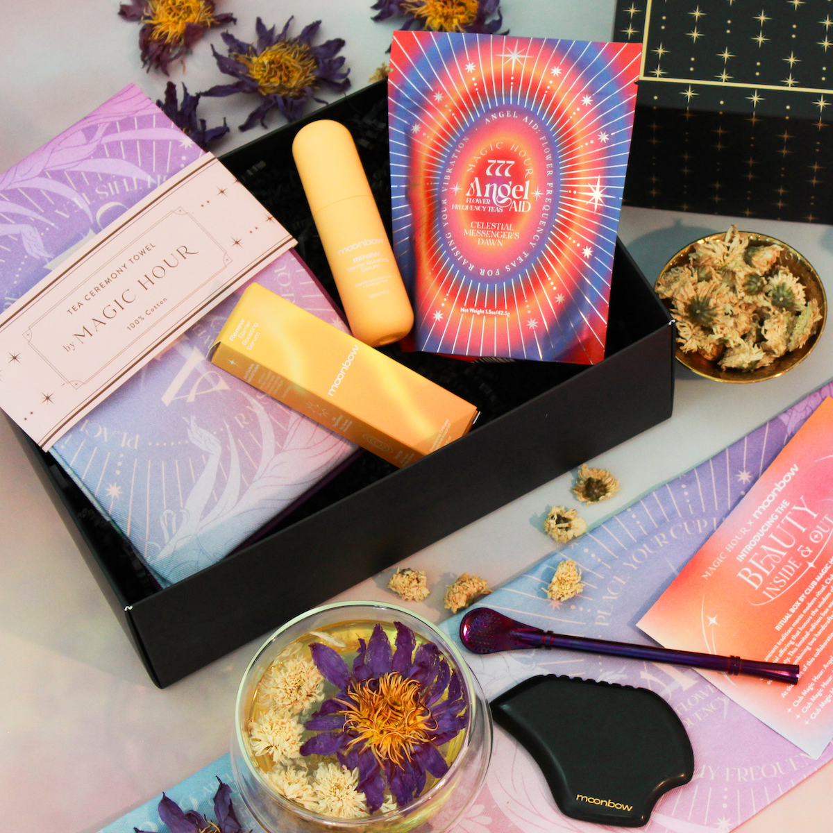 A flat lay of the Beauty Inside & Out Box featuring a botanical serum, cosmic-themed cards, dried flowers, and a black massage tool, all artfully arranged on a purple and peach backdrop.