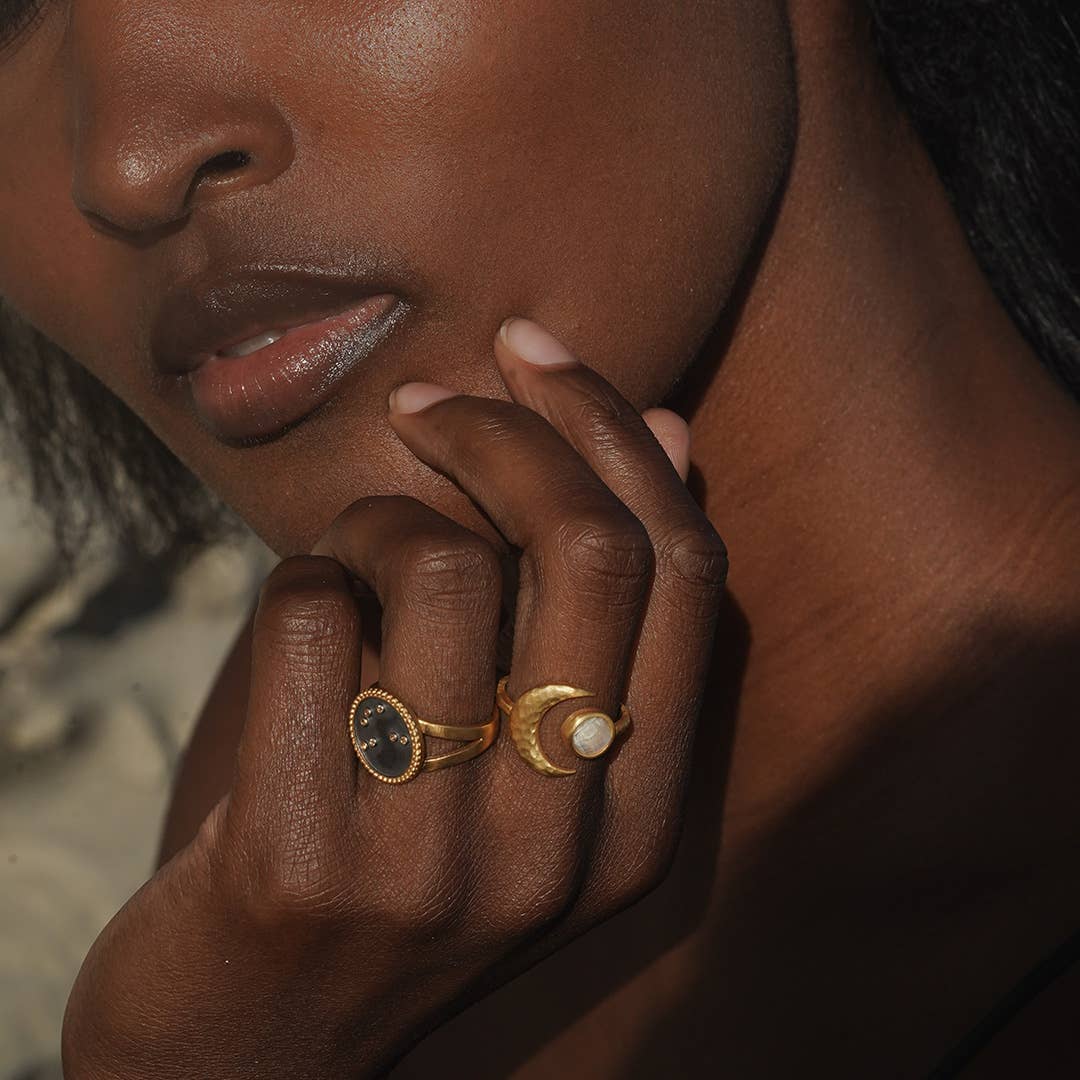 A person with dark skin gently touches their face, showcasing two gold rings—one with a dark, oval stone and the Cradled in Light Moonstone Ring—against a softly lit background.
