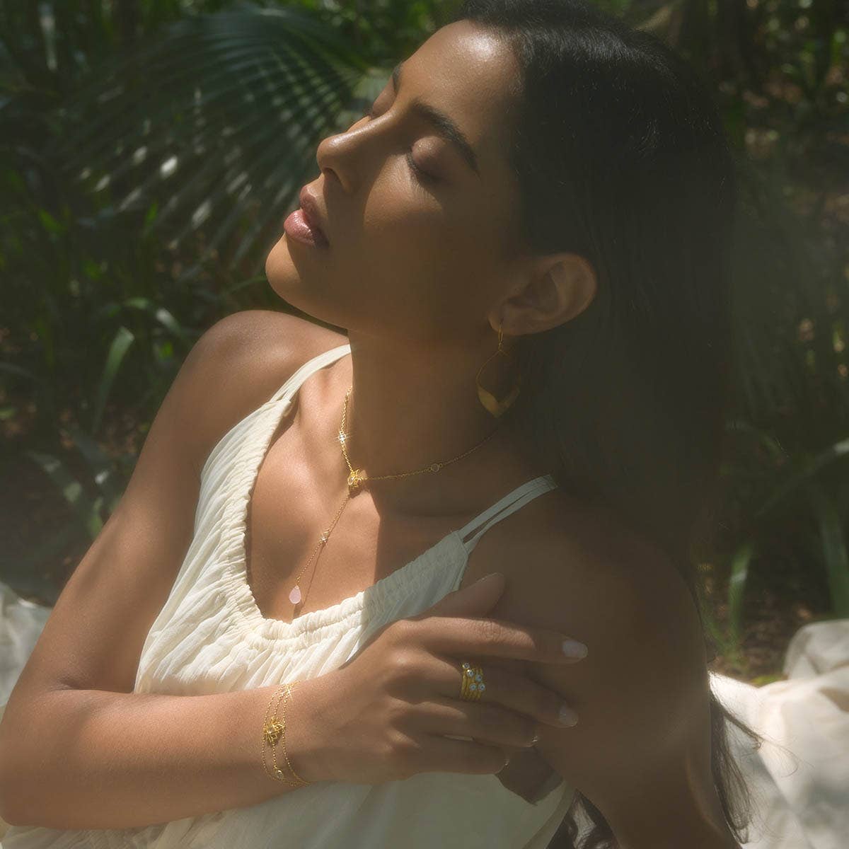 A woman in a cream dress sits outdoors, eyes closed in sunlight. She wears gold jewelry, including the Transformed by Love Lotus Lariat Necklace, with matching bracelet, ring, and earrings, gently resting her hand on her shoulder amid greenery.