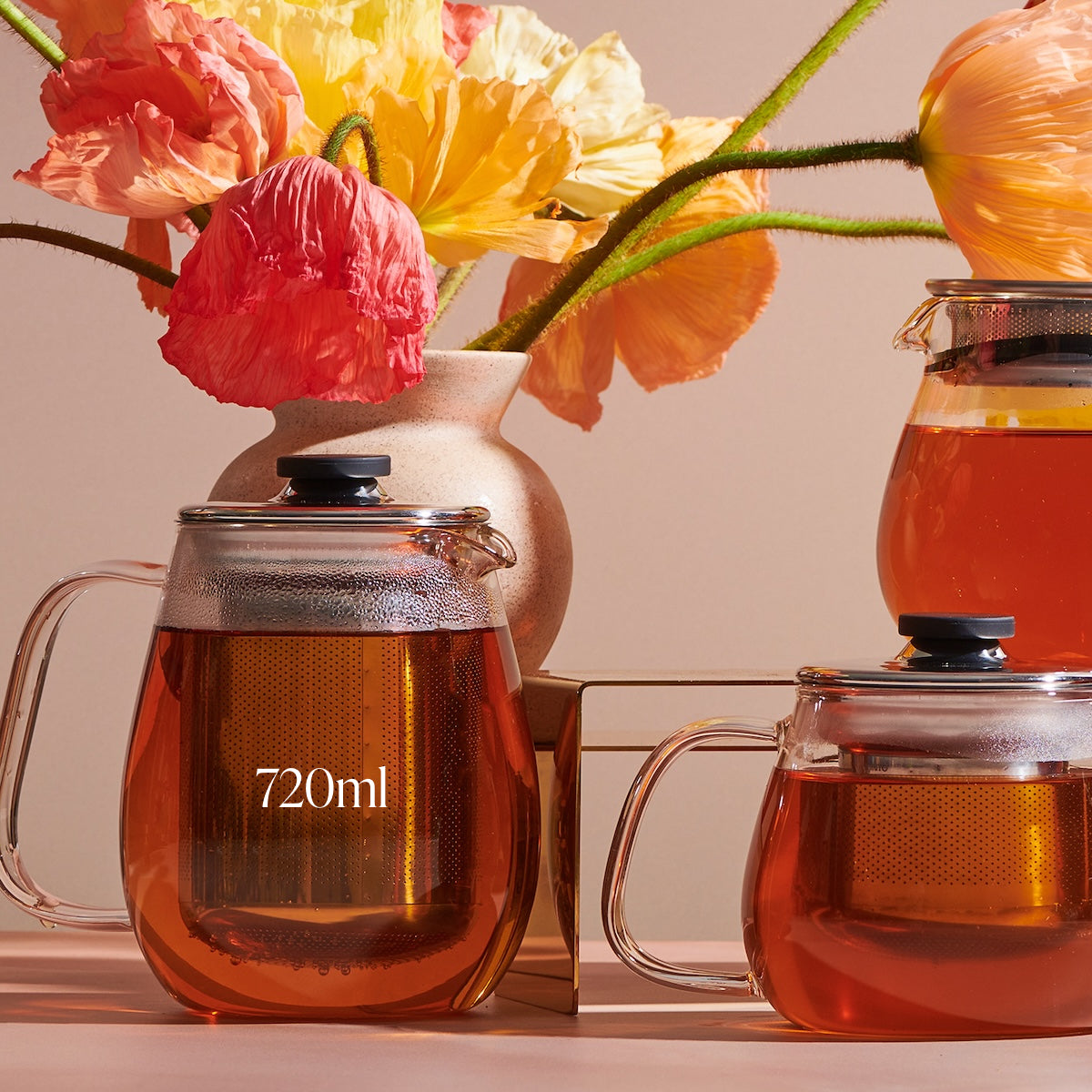 Glass teapots filled with amber-colored Magic Hour Kinto Stainless Steel Teapot are displayed. The largest teapot, labeled "720ml," sits on the left with another teapot to the right on a metal stand. Bright flowers in a vase are in the background, adding a colorful contrast.