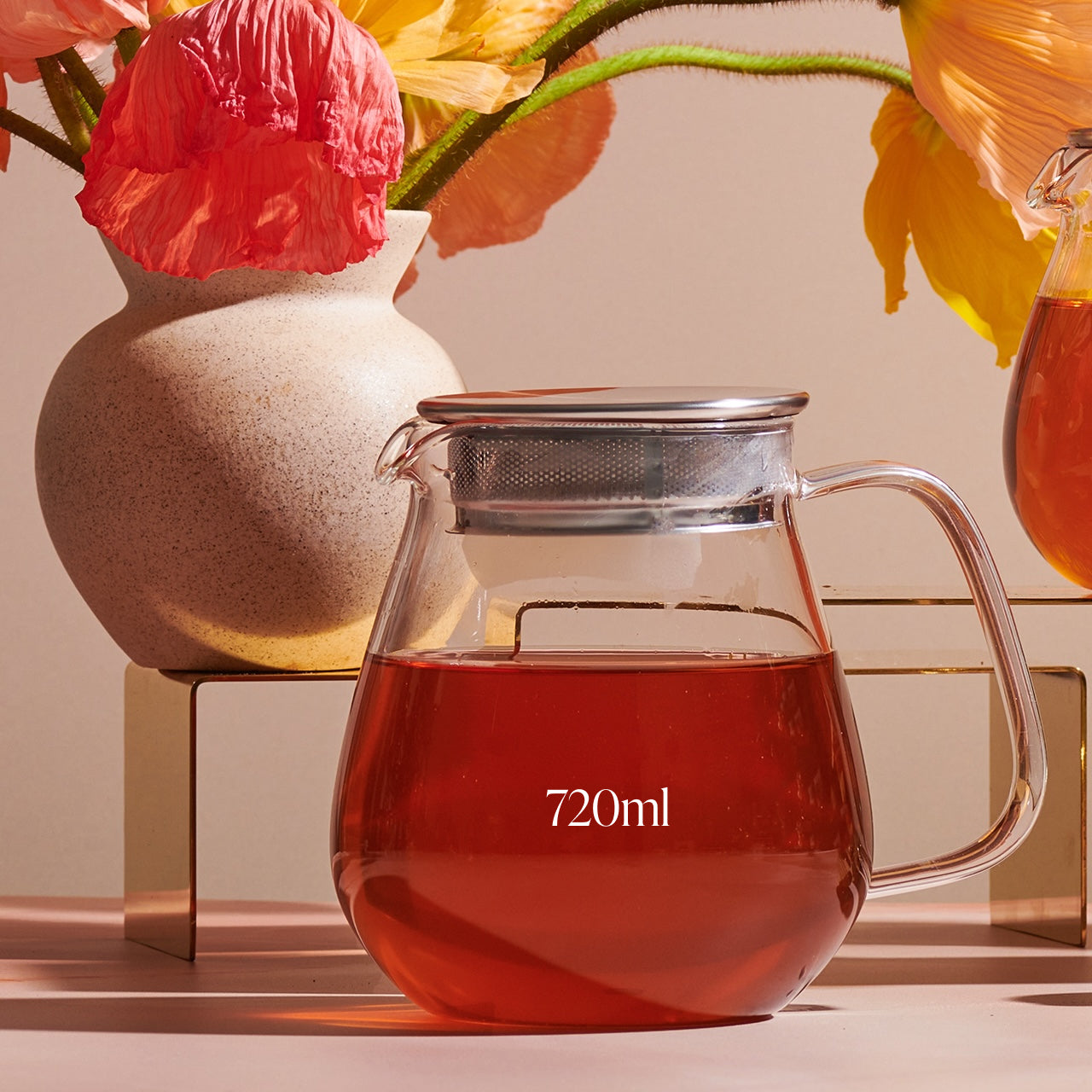 A Kinto One Touch Teapot marked "720ml" filled with red liquid, possibly Magic Hour Tea, sits on a table. Behind it, there is a ceramic vase holding vibrant pink and yellow flowers. The scene is well-lit with a soft, warm light.