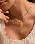 A woman wears a beige ribbed tank top and three gold necklaces, including the Abundant Grounding Tree Necklace with round pendants, plus another necklace with small colored beads. Her hand gently touches her chin.