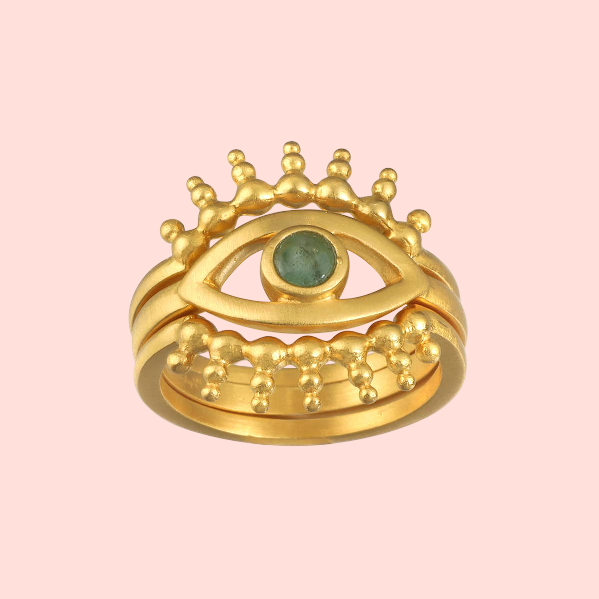 The Emerald Radial Evil Eye Ring set of 3 features gold-plated eye-shaped rings with green gemstone irises and gold bead accents resembling eyelashes, displayed against a pale pink background.