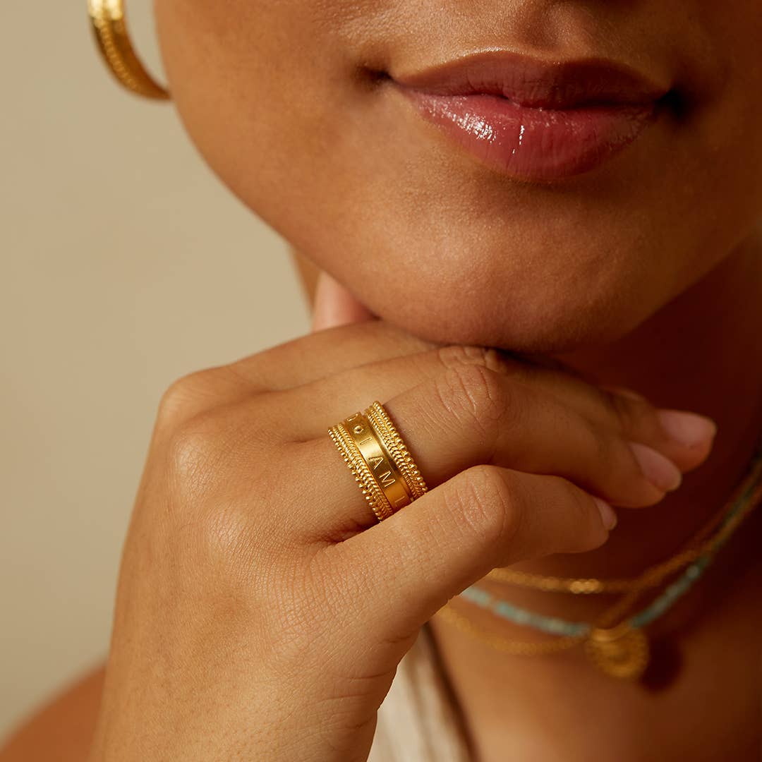 A close-up of a woman's face and hand shows her wearing the Inner Light I Am Loved Spinning Ring, paired with gold plated hoops and layered necklaces. Her lips are slightly parted as she smiles naturally, resting her chin on her hand.