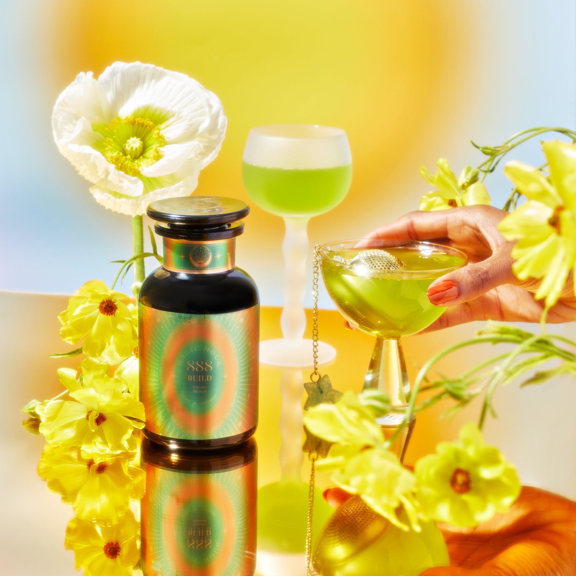 A vibrant arrangement features a black bottle labeled &quot;Ssss&quot; with white and yellow flowers. A hand holds a green School of Magic Tea in a stemmed glass, while another green drink sits in the background, all set against a bright backdrop for this mindfulness journey.