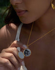A woman in a white top holds the Take Flight Butterfly Pearl Locket Necklace 18” with a black-and-white photo inside. She also wears a gold leaf earring. The background is green and softly blurred.