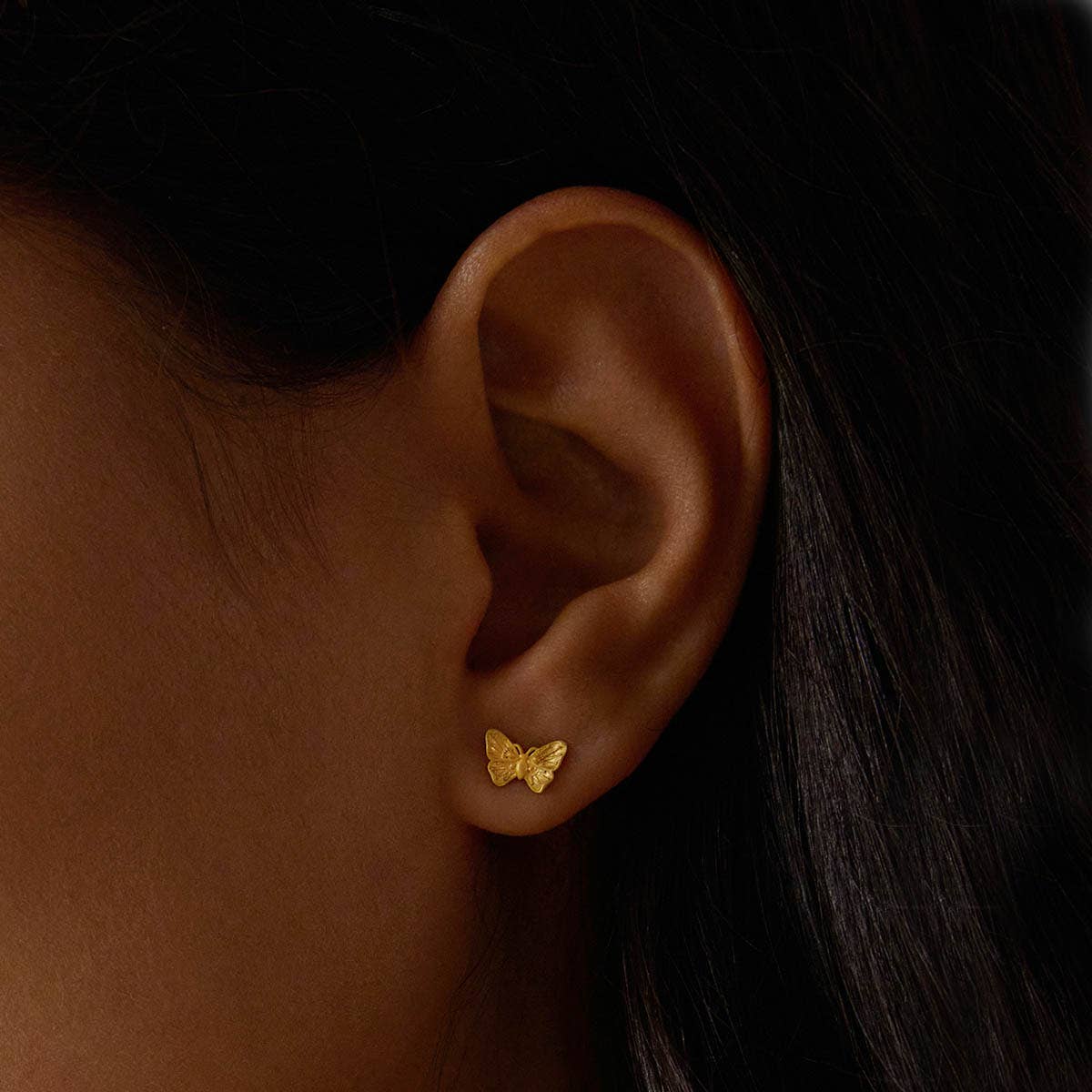 Close-up of a person’s ear wearing Freedom to Soar Butterfly Stud Earrings in 18k gold plate. The dark background highlights the jewelry’s transformative design alongside the person’s dark hair and skin.