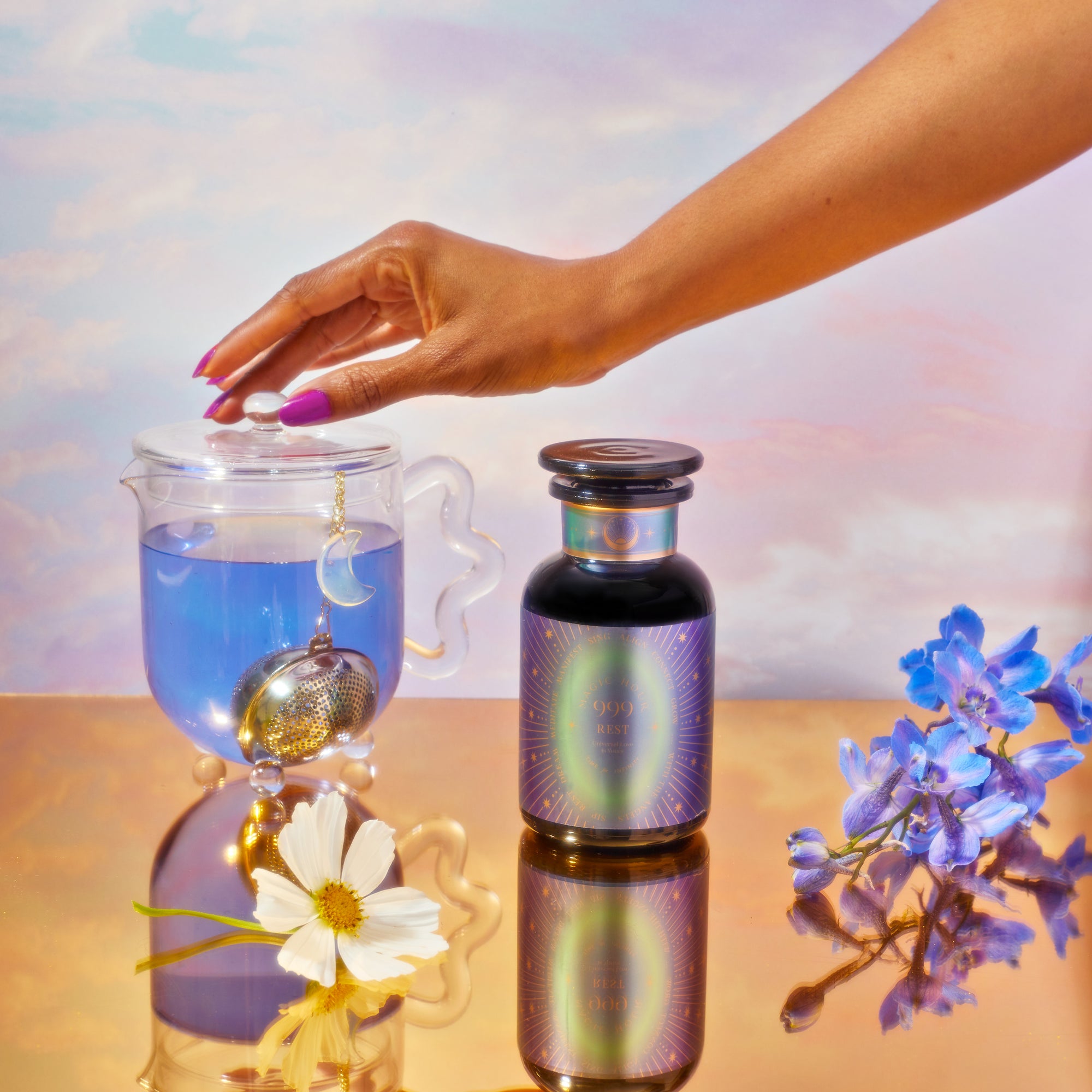 A hand with pink nails holds a glass teapot with blue wellness tea and a crescent moon infuser, beside a School of Magic Tea Subscription Box - Petite Apothecary Jar. Blue blossoms and a white flower surround them on a reflective golden surface against a pastel sky.