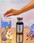 A hand with pink nails holds a glass teapot with blue wellness tea and a crescent moon infuser, beside a School of Magic Tea Subscription Box - Petite Apothecary Jar. Blue blossoms and a white flower surround them on a reflective golden surface against a pastel sky.