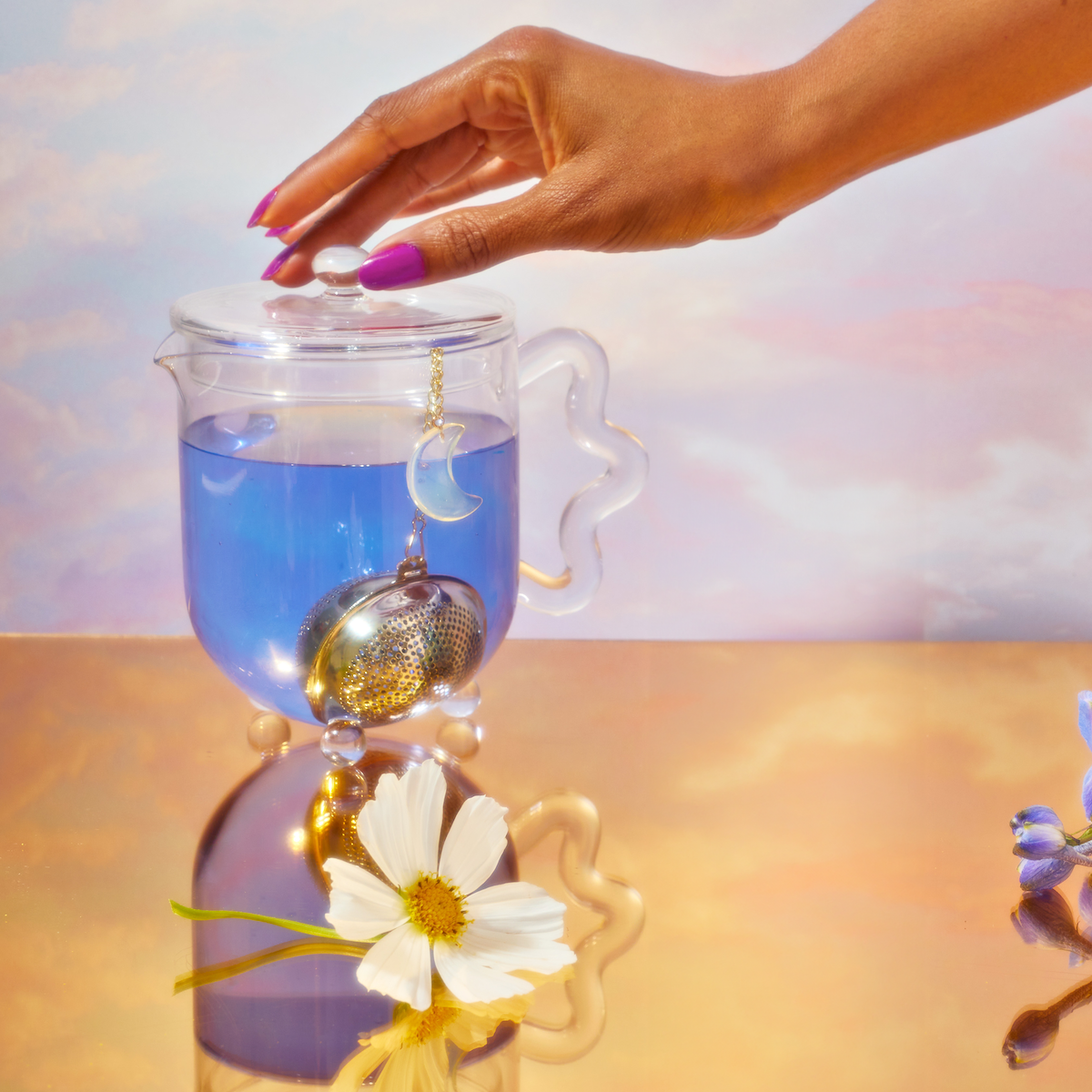 A hand rests on a glass teapot lid holding blue tea from the School of Magic Tea Subscription Box - Traveler Jar. A crescent moon infuser is submerged, and a white daisy graces the reflective golden surface, creating a spiritual connection amid soft pastels in the background.