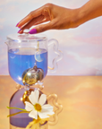 A hand rests on a glass teapot lid holding blue tea from the School of Magic Tea Subscription Box - Traveler Jar. A crescent moon infuser is submerged, and a white daisy graces the reflective golden surface, creating a spiritual connection amid soft pastels in the background.