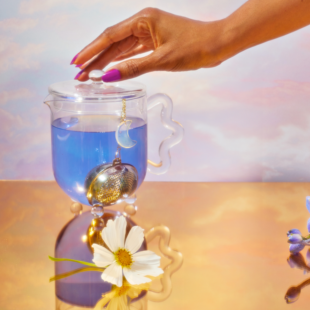 A hand lifts the lid of a glass teapot filled with 999: Divine Completion tea. A moon-shaped infuser hangs inside, while a white flower rests on a reflective gold surface against a pastel sky backdrop.
