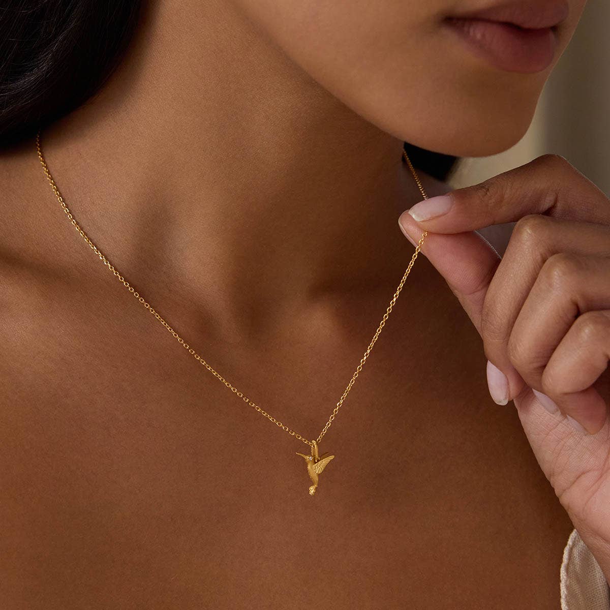 A woman holds the Loving Devotion Hummingbird Necklace 16", a gold pendant with shimmering white topaz, resting closely against her collarbone.