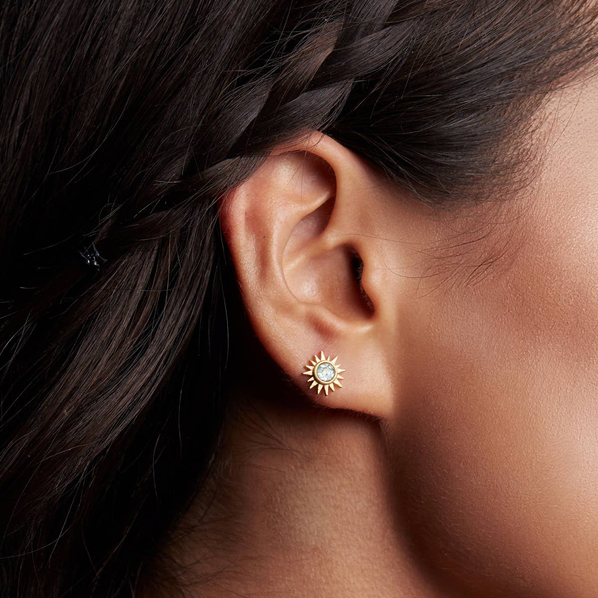 A close-up of a woman with braided dark hair wearing Blue Topaz Starburst Earrings—sun-shaped studs with blue topaz centers set in 18k gold plate.