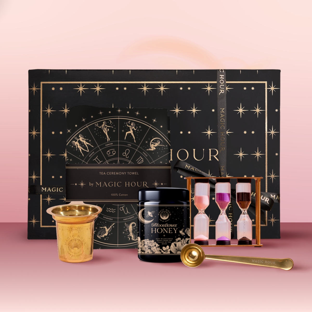 The Everything But The Tea Gift Set includes a starry black box, moon-themed tea towel, artisan tea strainer, Moonflower honey jar, gold spoon, and a sand timer with pink, white, and purple sands on a pink background.