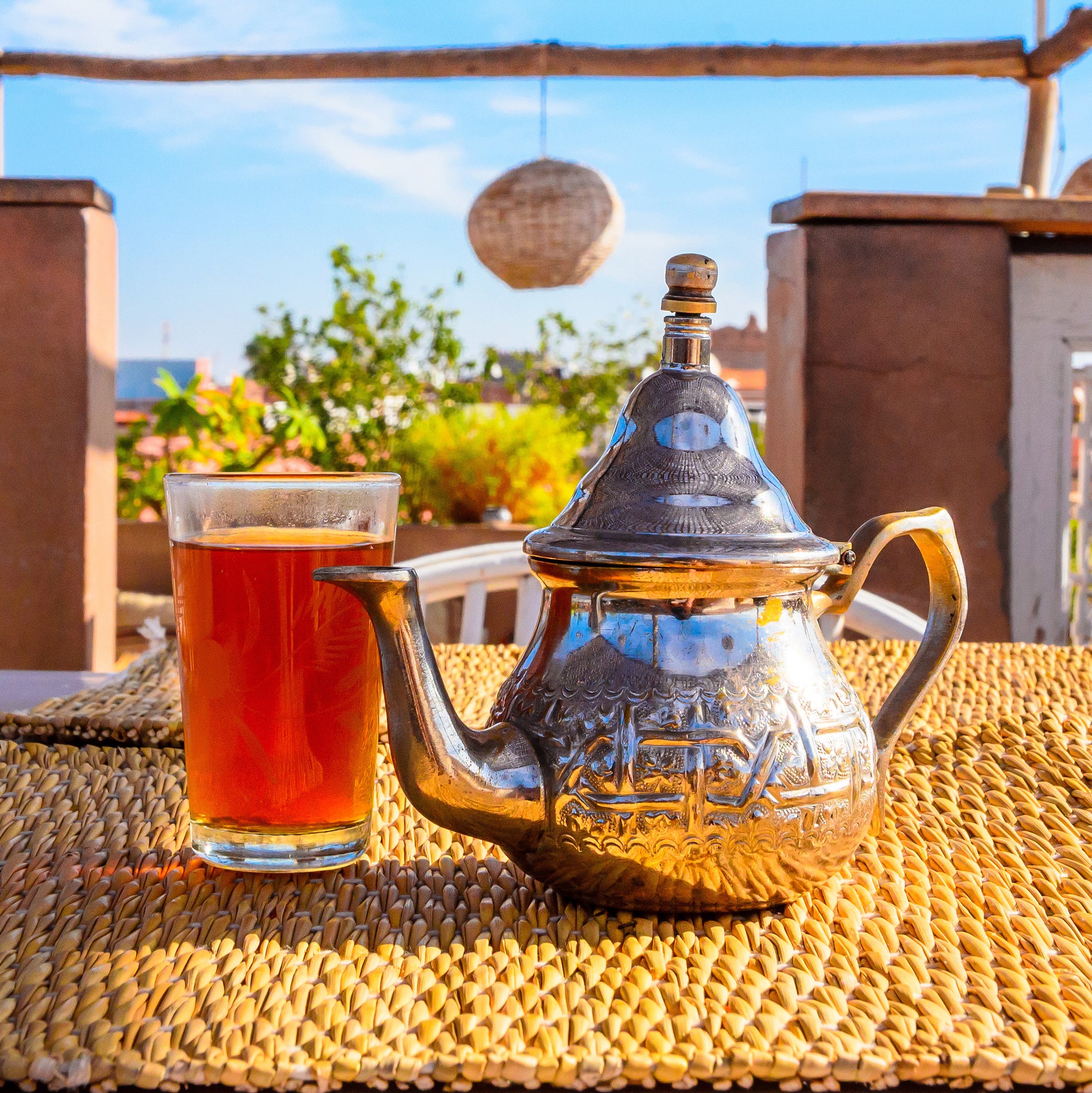 A glass of tea and a decorative silver teapot are placed on a woven placemat. The table is set outdoors with a sunny sky and green plants in the background, creating a relaxed and pleasant atmosphere.