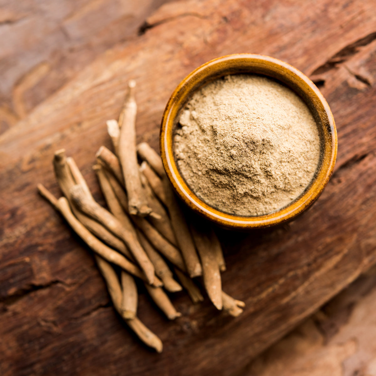 A small wooden bowl filled with ashwagandha powder is placed on a rustic wooden surface. Next to the bowl, several whole ashwagandha roots are scattered, showcasing the raw and powdered forms of the herb.