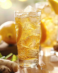 A refreshing glass of Magic Hour's Cream Soda White Tea: Ceremonial Soda Tea with Sugar Destroyer, filled with visible ice cubes and lemon slices. In the background, there's another similar drink accompanied by various ingredients including ginger root, lemons, and a hint of white tea. The setting is bright and sunny, suggesting a warm day.