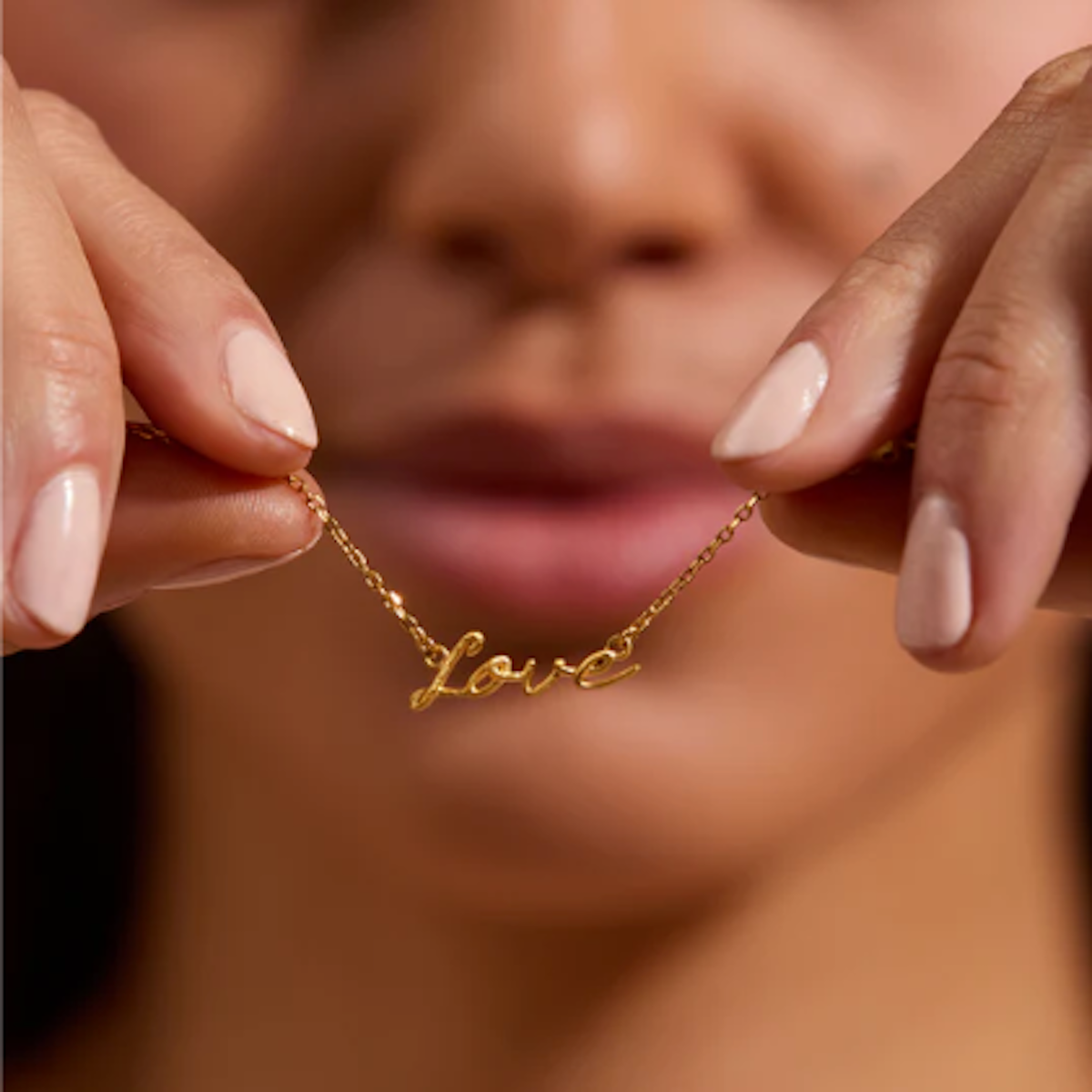 A person with light brown skin is holding up the Adorned with Love Gold Necklace, featuring 18kt gold plating and the word "love" written in delicate script. The background is blurred, drawing attention to the necklace that adds a personalized layered look. The person's nails are painted light pink.