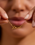 A person with light brown skin is holding up the Adorned with Love Gold Necklace, featuring 18kt gold plating and the word "love" written in delicate script. The background is blurred, drawing attention to the necklace that adds a personalized layered look. The person's nails are painted light pink.