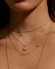 Close-up of a person's neck adorned with the "Adorned with Love Gold Necklace," an 18kt gold plated piece that adds elegance to a personalized layered look. The necklace features various pendants, including one in delicate script spelling "love," a floral design, and a small heart. The lighting casts soft shadows, highlighting the jewelry beautifully.