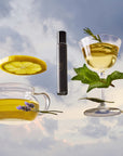 A glass teapot with herbal tea, a lavender sprig, lemon slice, fresh mint, rosemary, a glass of yellow drink, and the Air Essential Oil Perfume bottle float against a cloudy sky backdrop.