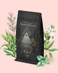 A black and gold tea package labeled "Magic Hour Air: Mint, Mullein & Citrus Tea for Respiratory Support" is surrounded by illustrated green leaves and white flowers on a pale pink background, highlighting its decaf green tea blend crafted to support respiratory health.